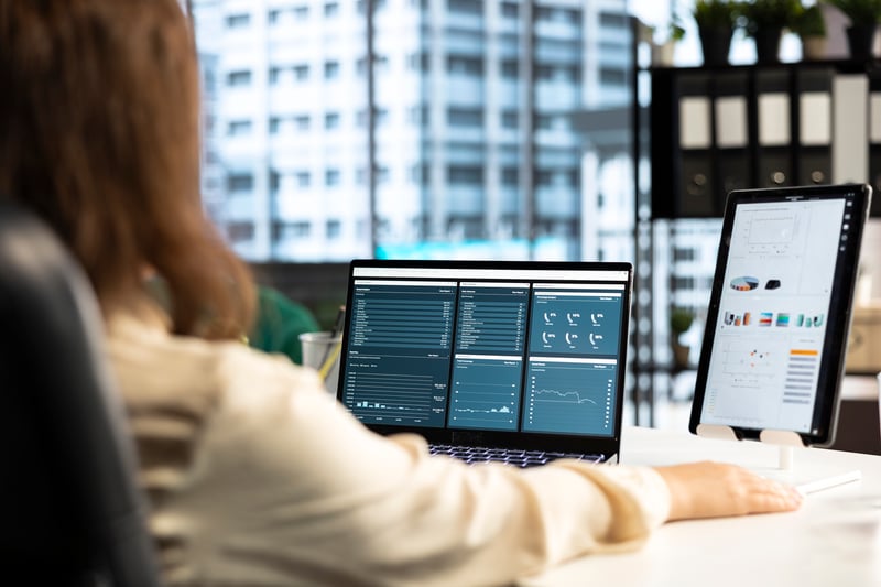 Vista desde atrás de una persona trabajando en una oficina frente a una laptop y una tablet que muestran dashboards con gráficos de rendimiento y datos durante las pruebas de sistemas de software