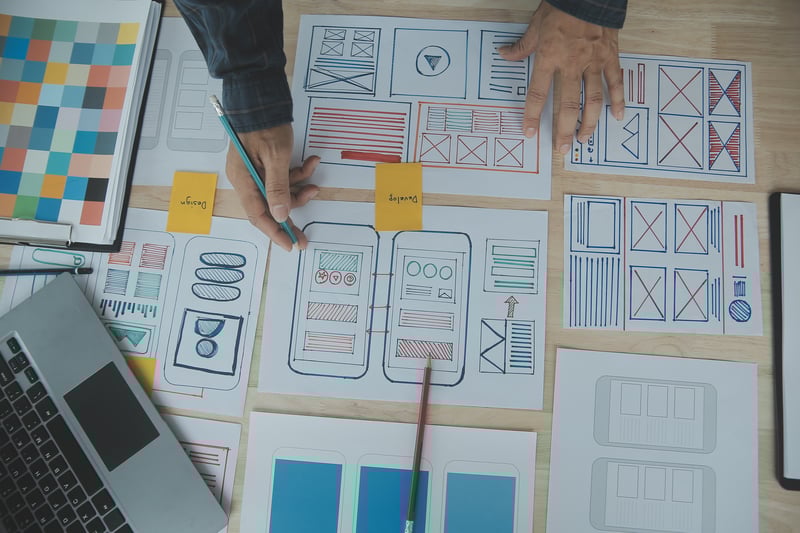 Vista superior de manos trabajando sobre bocetos de baja fidelidad y wireframes de una aplicación móvil en papel, utilizados para planificar y ejecutar pruebas de usabilidad en las etapas tempranas del diseño UX